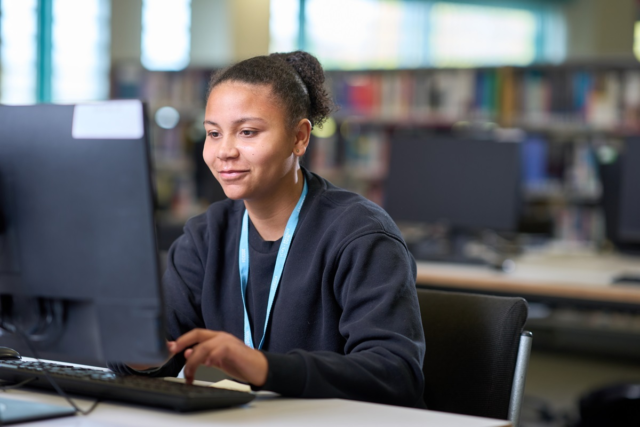 Student working at a computer in the learning centre with computer desks and bookshelves behind her